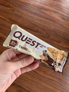 a hand holding a bar of quest peanut butter sandwich on a wooden table with wood grain flooring