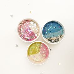 three bowls filled with different colored glitters on top of a white table next to stars