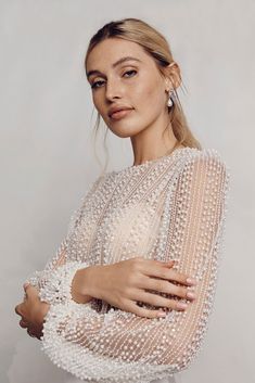 a woman with her arms crossed, wearing a white dress and pearls on the sleeves