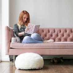 a woman sitting on a pink couch reading a book with a white pouffle