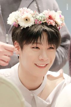 a young man with flowers in his hair smiles at the camera while another person stands behind him