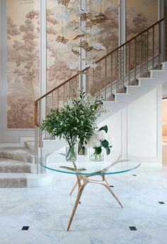 a glass table with flowers on it in front of a stair case and wallpaper