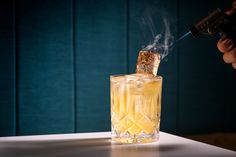 a person holding a lighter over a glass filled with ice and lemonade on top of a table