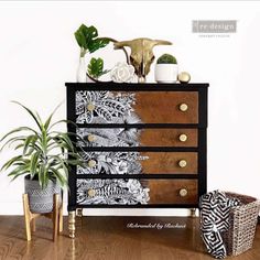 a black and white chest of drawers next to two plants on top of a wooden floor