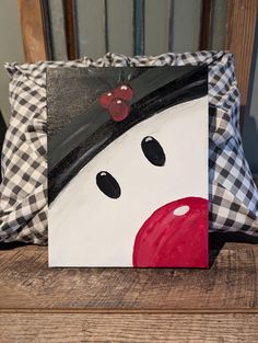 a painting of a snowman on a wooden chair next to a plaid pillow with a red nose