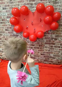 a young boy holding a stick in front of a red heart shaped balloon wall hanging on a brick wall