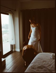 a woman in a white dress standing next to a bed and another person kneeling down