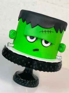 a green cake with black trim and eyes on it's head, sitting on top of a pedestal