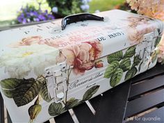 an old suitcase with flowers painted on it sitting on a bench in front of some flowers