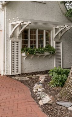 a brick walkway leading to a white house with windows and plants in the window boxes