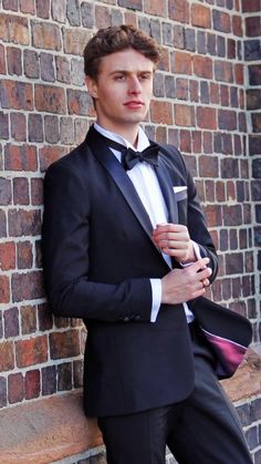 a man in a tuxedo leaning against a brick wall with his hand on his pocket