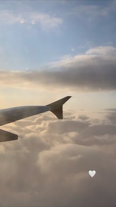 the wing of an airplane flying in the sky with clouds and a heart shaped object