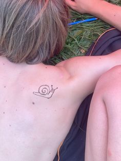 a woman laying on the grass with her back turned to the camera while she has a snail tattoo on her left shoulder