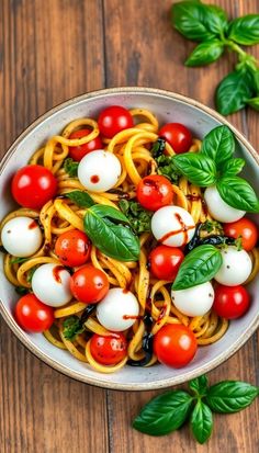 a bowl filled with pasta, tomatoes and basil on top of a wooden table next to green leaves