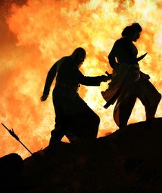 two people holding hands in front of a large fire that is blazing up into the sky