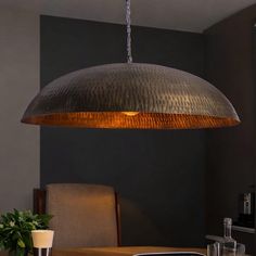 a large metal light hanging from the ceiling over a dining room table with chairs and a potted plant