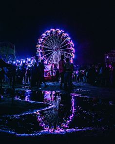 people are standing around in front of a ferris wheel at night with bright lights on it