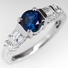 The lovely sapphire engagement ring is centered with a 1.23 carat round cut blue sapphire set into a four-prong head. The top of the shoulders is accented with two (2), channel set, baguette cut diamonds and one (1), prong set, round brilliant cut diamond on each side. The top and sides of the shank feature engraved wheat motifs. The ring measures 6.0mm at the top, rises 7.1mm above the finger, tapering to 1.9mm wide and 1.7mm thick at the base of the shank. This ring is currently a size 6.75 an Blue Sapphire Engagement Ring, Blue Engagement Ring, Round Sapphire, Sapphire Engagement Ring Blue, Sapphire Engagement Ring, Baguette Cut Diamond, Emerald Jewelry, Channel Set, Baguette Cut