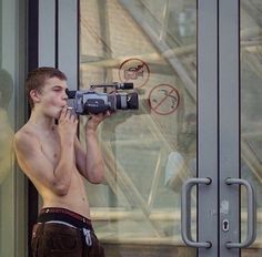 a shirtless man holding a camera up to his face while standing in front of a building