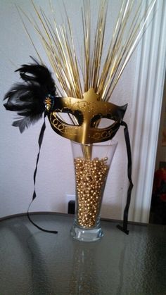 a masquerade mask sitting on top of a glass vase filled with gold and black feathers