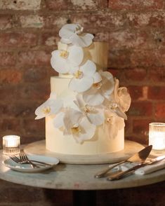 a three tiered cake with white flowers and candles on a table in front of a brick wall