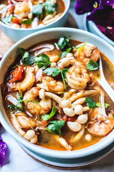 two bowls of soup with shrimp, beans and basil on the side next to purple flowers