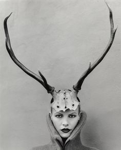 a woman wearing a deer's head mask with horns on her head, in front of a wall