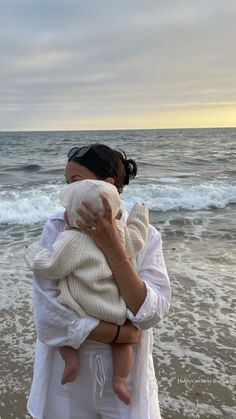 a woman holding a baby in her arms while standing on top of a beach next to the ocean