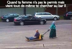 a woman pushing a man on a lawn mower in front of some parked cars