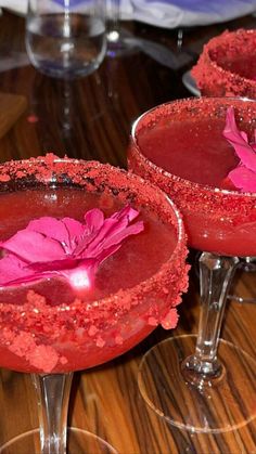 two wine glasses filled with red liquid and pink flowers on top of a wooden table