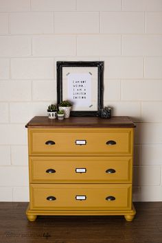 a yellow dresser with drawers in front of a white brick wall and framed photograph on top