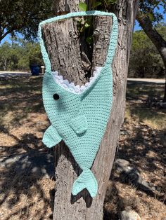 a crocheted blue fish hanging on a tree in front of some trees and grass