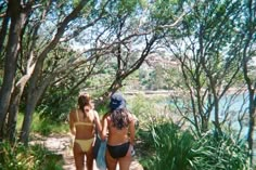 two women in bikinis walking down a path next to trees and water on a sunny day