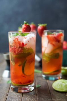 two glasses filled with fruit and ice on top of a wooden table next to limes