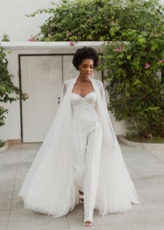 a woman in a white wedding gown and cape walking down the street with her hands on her hips