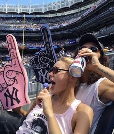 two women sitting in the stands at a baseball game, one holding up a foam finger sign