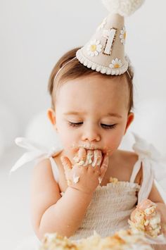 a baby girl wearing a party hat eating cake with her hands and face covered in frosting