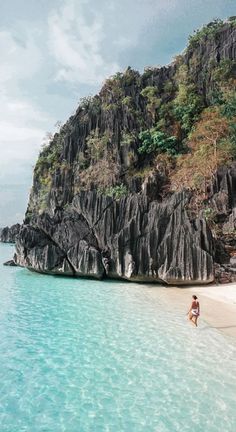 a person walking on the beach in front of a rock formation and clear blue water