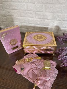 two pink and gold jewelry boxes sitting on top of a wooden table next to purple flowers