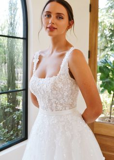 a woman in a wedding dress standing by a window with her hands on her hips