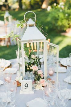 a table topped with a white lantern filled with flowers and greenery next to glasses