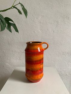 an orange vase sitting on top of a white table next to a green leafy plant