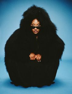 a woman in a black fur coat sitting on top of a blue floor with her hands on her knees