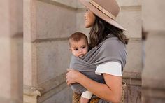 a woman carrying a baby in a grey and tan wrap on her back while standing next to a building