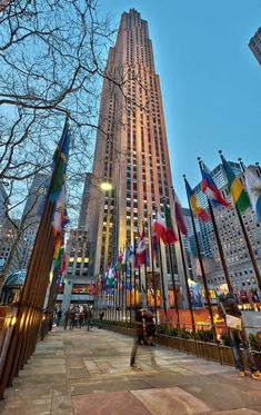 the tall building has many flags in front of it and people are walking on the sidewalk