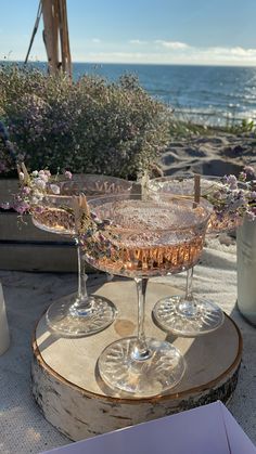 two wine glasses sitting on top of a wooden table next to the ocean and lavender flowers