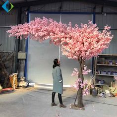 a woman standing next to a tree with pink flowers on it in front of a wall