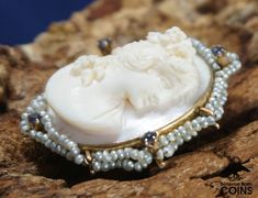 "Metal: 10k Yellow Gold (.417) Weight: 8.4 grams Measurements: 1.25\" Long; 1\" Wide; 0.54\" Deep Markings: \"10k\" *This stunning brooch features a cameo of a finely dressed woman looking off to the right hand-carved on a pink/white shell, set into a 10k yellow gold border and bezel. The border features small pearls in two criss-crossed rows, with 4 small purple stones. The brooch features impressive high relief carving. The brooch also functions as a necklace pendant. This outstanding brooch i White Oval Brooch For Anniversary, Heirloom White Brooch Jewelry, White Heirloom Brooch Jewelry, Oval Gemstone Brooches For Weddings, Wedding Pendant Brooches With Cabochon, Wedding Brooches With Cabochon Pendant, Wedding Brooch With Cabochon Pendant, Victorian White Cabochon Brooches, Victorian Style White Cameo Brooch