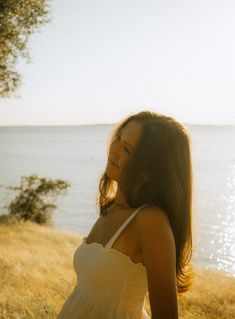 a woman in a white dress standing by the water with her hair blowing in the wind