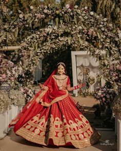 a woman in a red and gold lehenga standing under an archway with flowers